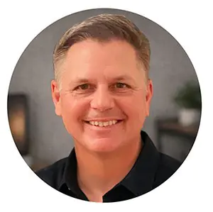 A professional headshot of Corey Gasman, a web designer, smiling and wearing a grey polo shirt against a neutral, light-colored background.