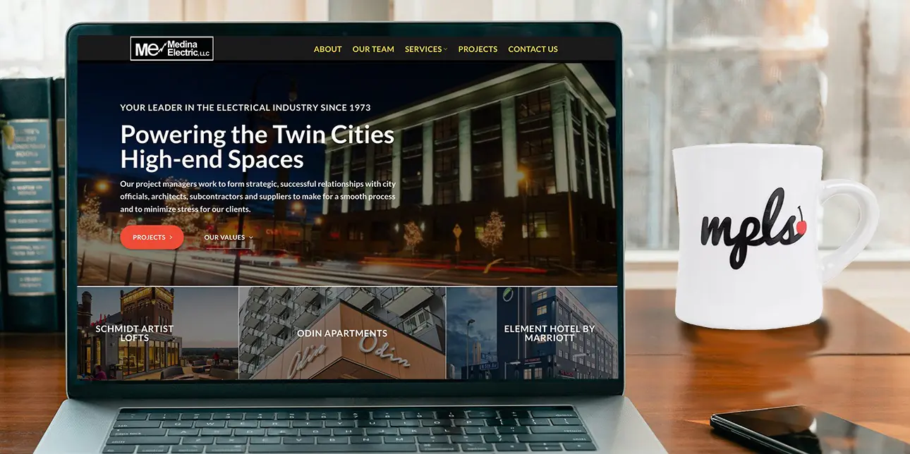 A professional over-the-shoulder view of a laptop screen displaying a custom WordPress website design, featuring a "Powering the Twin Cities Spaces" headline and a minimalist layout, set against a blurred indoor background.