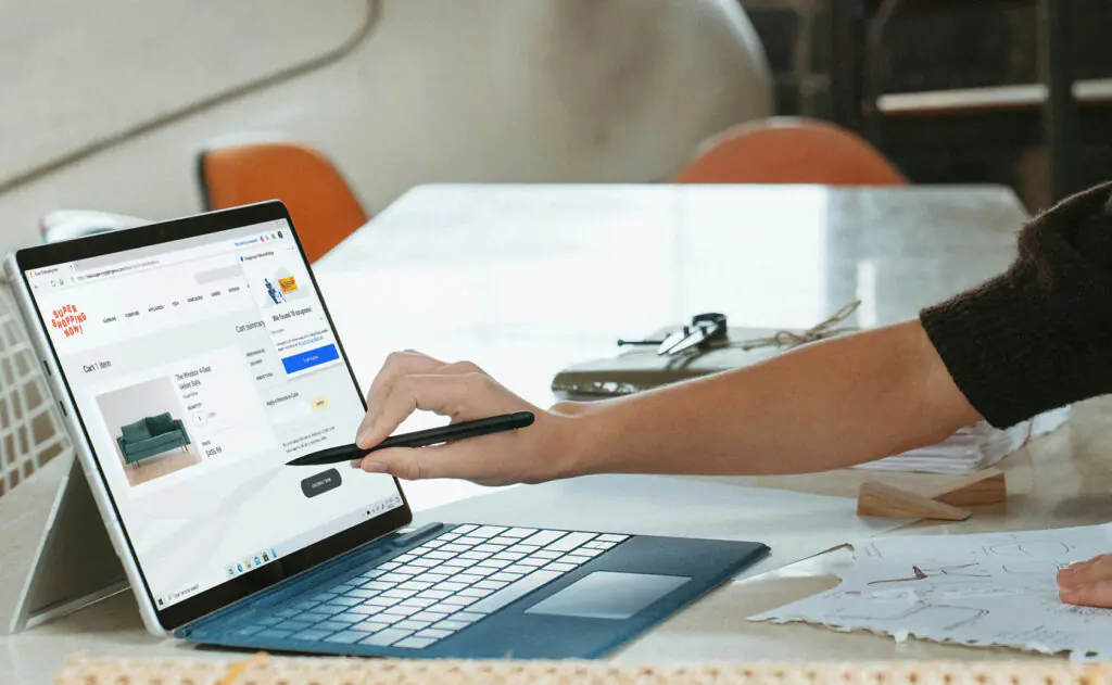 A professional over-the-shoulder view of a person using a laptop and tablet at a wooden desk to design a small business website, featuring clean layouts and modern interface elements.